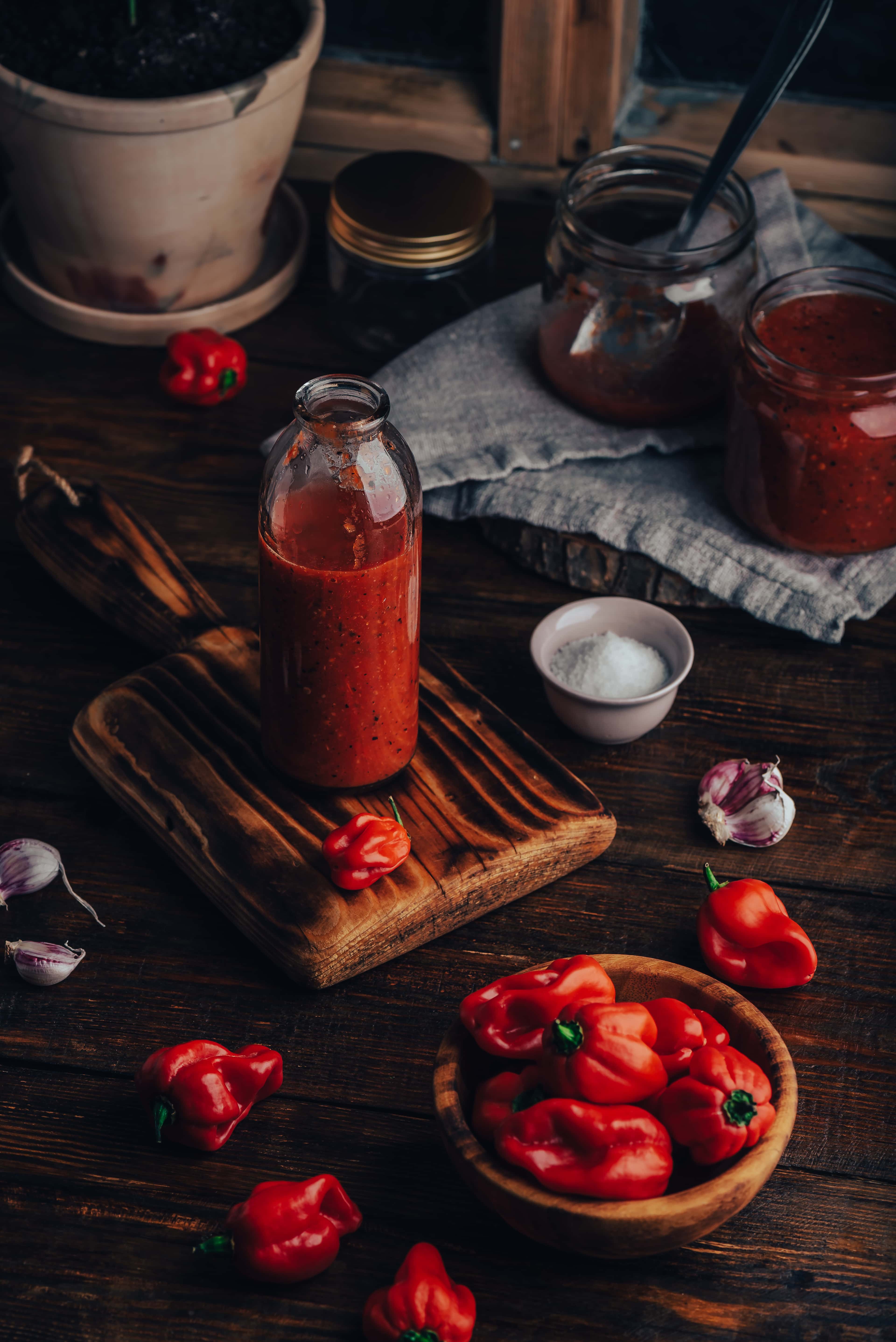 Glass bottle of red hot sauce on a dark wooden board with salt, garlic, and fresh red peppers